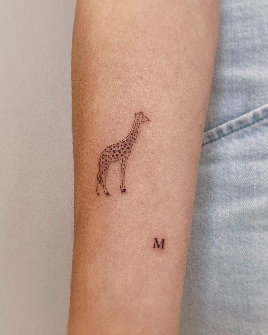 Una jirafa y una pequeña M tatuadas en el brazo